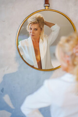 a middle-aged woman in a white jacket topless looks at reflection in a mirror.