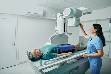 Male patient lying on bed while female nurse adjusting modern X-ray machine for scanning his leg or...