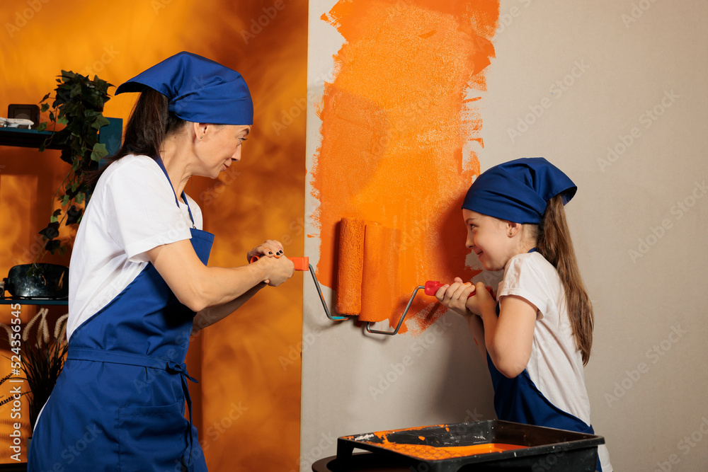 Wall mural Young people using orange color paint, renovating apartment walls with painting tools or equipment. Mom and child redecorating house room with roller and paintbrush, having fun.