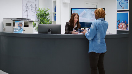 Female patient paying consultation with credit card at reception desk, attending checkup visit appointment. Woman talking to receptionist to pay medical consultation at clinical counter. - Powered by Adobe