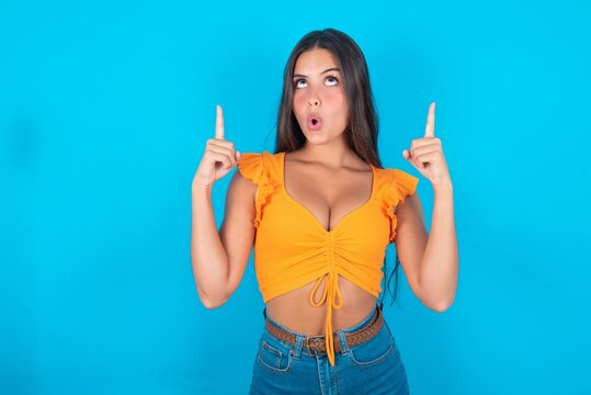 beautiful brunette woman wearing orange tank top over blue background being amazed and surprised looking and pointing up with fingers showing something strange.