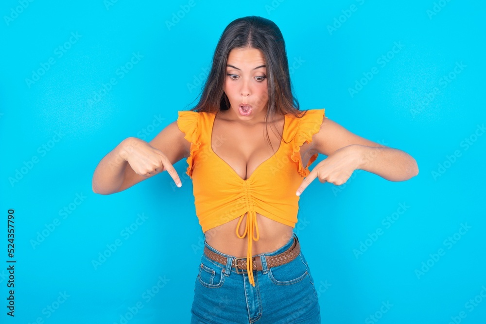 Wall mural amazed brunette woman wearing orange tank top over blue background points down with fore fingers, op