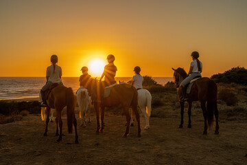 6 students of a riding school riding their horses watching the sunset near the sea.