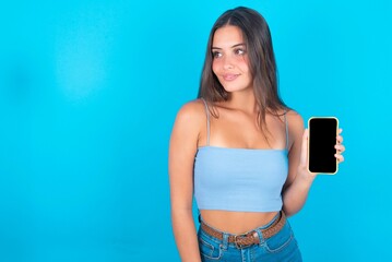 beautiful brunette woman wearing blue tank top over blue background holds new mobile phone and looks mysterious aside shows blank display of modern cellular