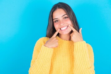 Happy beautiful brunette woman wearing yellow sweater over blue background with toothy smile, keeps index fingers near mouth, fingers pointing and forcing cheerful smile - Powered by Adobe