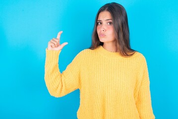 Displeased beautiful brunette woman wearing yellow sweater over blue background shapes little hand sign demonstrates something not very big. Body language concept.