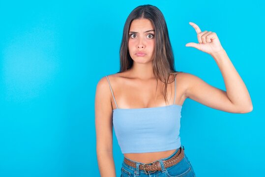beautiful brunette woman wearing blue tank top over blue background purses lip and gestures with hand, shows something very little.