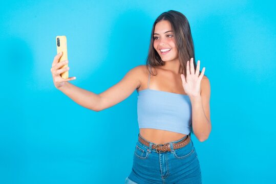 beautiful brunette woman wearing blue tank top over blue background holds modern mobile phone and makes video call waves palm in hello gesture. People modern technology concept