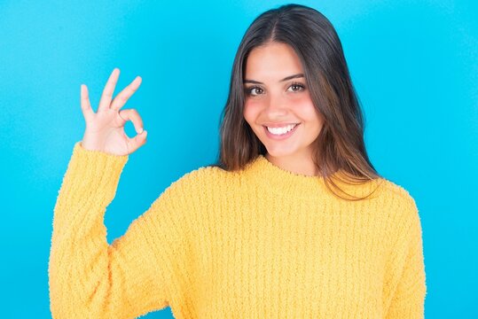 beautiful brunette woman wearing yellow sweater over blue background hold hand arm okey symbol toothy approve advising novelty news