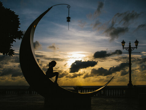 Girl sits on a moon statue with sunset and ocean behind her