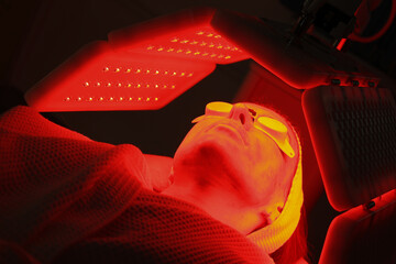 Express facial treatment with led therapy. Beautiful girl on a light therapy procedure. LED lamp with red and yellow light. Safe skin care. Woman in protective glasses. Beauty and wellness concept. 