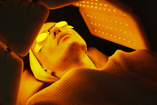 Express facial treatment with led therapy. Beautiful girl on a light therapy procedure. LED lamp with red and yellow light. Safe skin care. Woman in protective glasses. Beauty and wellness concept. 