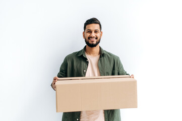 Positive attractive candid arabian or indian young delivery man in casual clothes, holding a cardboard box, standing on isolated white background, looks at camera, smiles friendly. Delivery concept