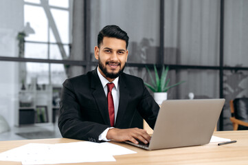 Portrait of attractive elegant confident arabian or indian successful businessman, entrepreneur, lawyer, in formal suit, sit at work desk with laptop in modern creative office, looks at camera, smile