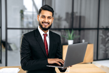 Fototapeta premium Portrait of successful confident elegant Indian or Arabian young businessman, in a suit, holding an open laptop in his hand, stand near the desktop in his modern office, looking at the camera, smiling