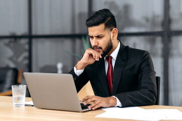 Confused puzzled indian or arabian young businessman, entrepreneur, in a suit, sitting in his modern office, focused look at laptop screen, see unexpected message or news, is under stress