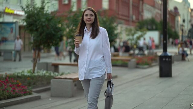 Modern businesswoman with a cup of coffee and a laptop in her hands walks along the sidewalk on the street. Cryptanalyst goes to the office to work with cryptocurrency
