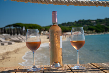 Glasses and bottle of cold rose wine from Provence served outdoor on wooden yacht pier with view on blue water and white sandy beach Plage de Pampelonne near Saint-Tropez, France