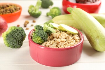 Feeding bowl with oatmeal porridge and vegetables on white wooden table, closeup. Natural pet food