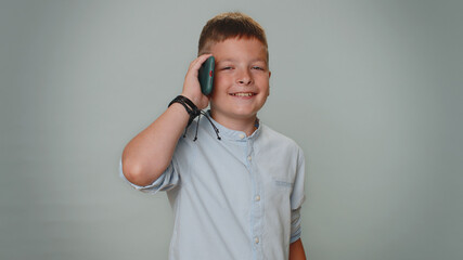 Toddler kid boy in shirt having pleasant mobile conversation using smartphone with friends or family, telling good news. Young teenager child schoolboy isolated alone on gray studio background indoors