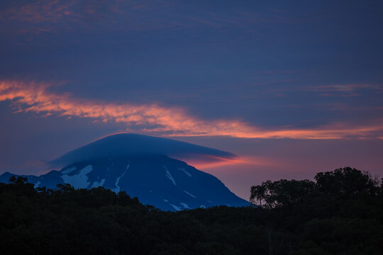Ilyinsky volcano (Ilyinskaya sopka) and cloud Altocumulus lenticularis A snow-capped Kamchatka volcano at dusk, partially covered by a lenticular cloud, with dramatic twilight skies.