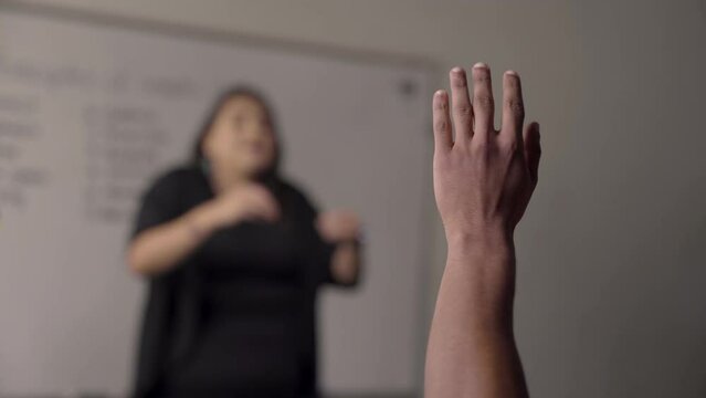 Medium frame of a Native American student raising their hand during class while the teacher lectures. Handheld.
