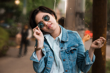 Pretty and cheerful young generation z girl with short hair dressed in blue with a denim jacket, sunglasses and gold jewelry posing in front of a reflective surface with a slight gold color.
