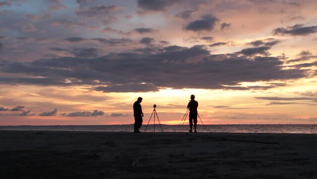 Silhouette of two photographers walking through the beach photographing a majestic view of sunset