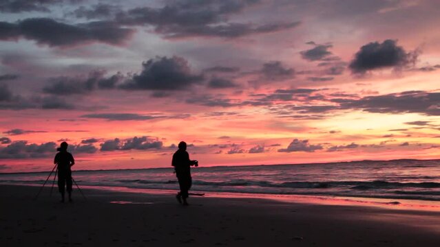 Silhouette of two photographers walking through the beach photographing a majestic view of sunset