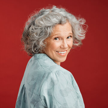 .. Confident, Happy And Smiling Senior Woman Feeling Playful And Cheerful While Posing Against Red Studio Background. Portrait Of A Beautiful, Older And Retired Lady Looking Back With Grey Hair.