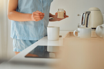 Fototapeta na wymiar Casual woman adding sugar while preparing a cup of coffee, tea or hot chocolate in the kitchen during the morning. Female hands holding teaspoon for a fresh beverage in a mug for breakfast at home