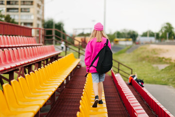 Fototapeta na wymiar A teenage girl with a backpack walks through the stands of the school stadium. Rear view