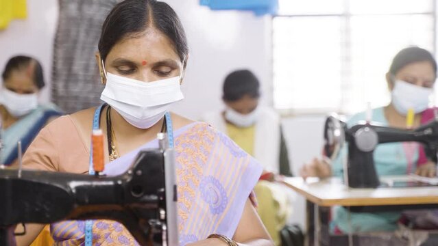 group of Women with medical face mask busy working on sewing machine at garments factory during coronavirus covid-19 pandemic - concept of pollution, safety and healthcare