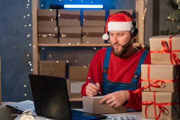 small business startup owner Santa Claus working at home with laptop. Online Christmas sale. man sits at a table with boxes and writes the delivery address. Holiday SME
