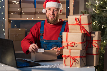Caucasian business owner working with product packaging and laptop wearing a Sanat Claus hat and...