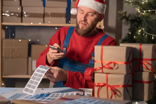 Young Santa Claus businessman sitting at his desk and scanning a delivery box with his mobile phone. Man selling products online on Christmas, scanning the barcode with his phone.
