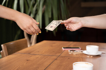Fototapeta na wymiar Woman hand holding US dollar banknotes giving cafe waitress, two cups of coffee on background. payment, service charge, bill checking, money tips