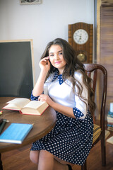 A cute teenage girl with long hair in a classic school dress in a classic interior with books and notebooks at the lesson. School life. Back to school