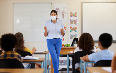 Young female teaching after covid pandemic, in classroom with young children students, in face mask. Back to school for little kids learning their education from their teacher after quarantine