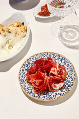 Meat antipasti on ceramic plate on white table with hard shadow. Meat platter with bresaola, hamon, prosciutto. Meat appetizer in Italian menu. Aesthetic food composition. Sunny day in tuscany.