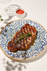 Grilled beef steak with rosemary on ceramic plate. Classic striploin steak cooked on grill. Bbq beef fillet. Steak in minimal style with shadows. Grilled striploin in summer menu. Meat dish on grill.
