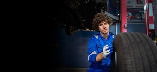 Fototapeta na wymiar A portrait of a young Caucasian mechanic fixing a tire at a garage at the auto mechanic service station. The mechanic inspects the tire. Auto repair service concept. banner image