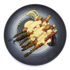 baked aspargus covered with bacon on a black ceramic plate isolated on white background
