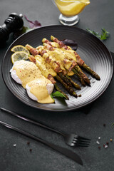 baked aspargus covered with bacon and poached eggs on a black ceramic plate