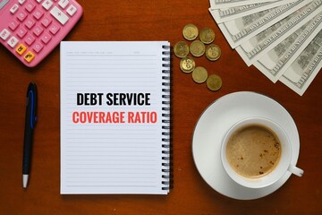 Flatlay picture of notebook written Debt Service Coverage Ratio, cup of coffee, calculator and money on the table.