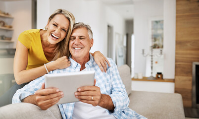 Mature couple laughing and smiling at funny app on digital tablet together, on their modern living room couch. Husband and wife sitting at home and looking at their touchscreen display device