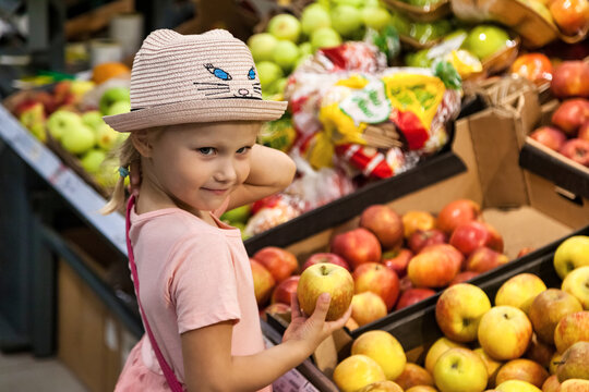 Portrait little girl with apple standing in grocery food fruit store, deep thinking and looking at camera. Adorable cute kid boy fresh fruits. Choosing healthy food concept. Copy space
