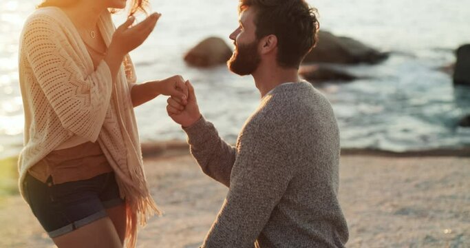 Romantic, romance and engagement proposal at the beach by boyfriend to surprise his girlfriend with a ring. Happy, in love and young man asking to marry a woman at sea on a summer evening sunset