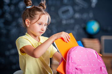 Cute happy schoolgirl puts stationery in a backpack. Preparation for school. Back to school. A little girl is learning to self-assemble a school backpack.