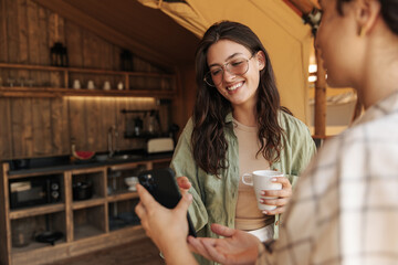 Nice young caucasian two women use smart phone spending time in camping during day. Brunette wears glasses, tank top and shirt. Online entertainment concept